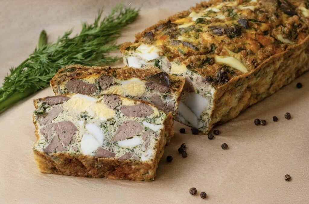 A classic French terrine, a staple among famous French foods, shown in a cross-section revealing layers of meat and egg, accompanied by fresh dill and black peppercorns scattered on a parchment background for a rustic culinary presentation.