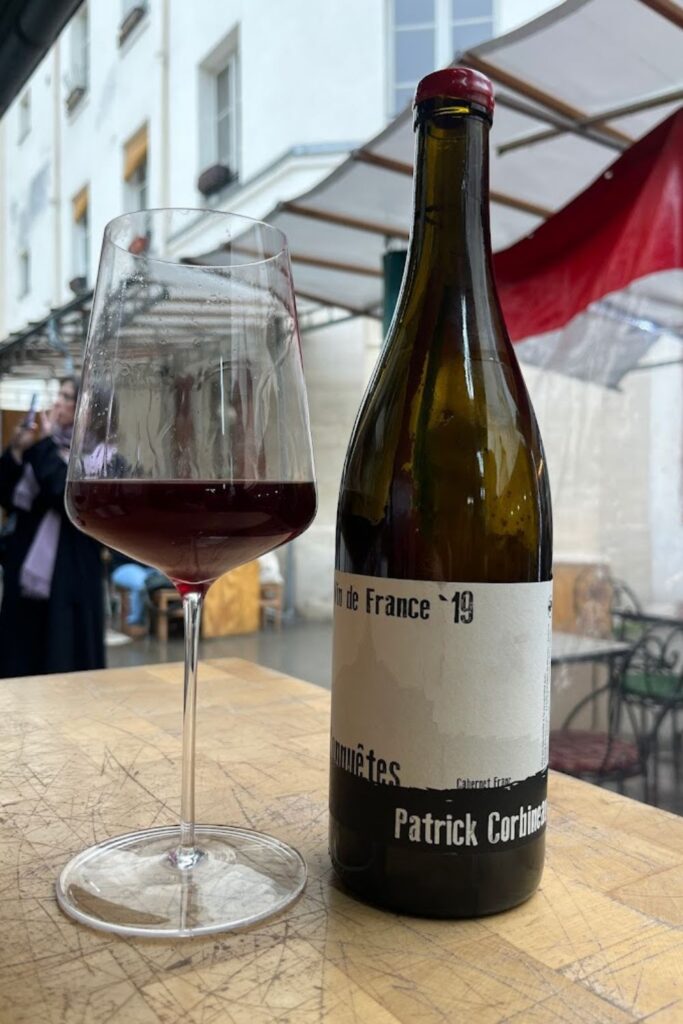 A glass of red wine next to a bottle of 'Vin de France '19 Communes' by Patrick Corbineau, positioned on an outdoor table, with the street life of Paris blurred in the background, a scene evoking the relaxed pleasure of the city's best wine bars.