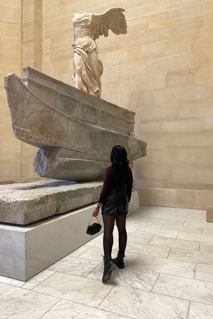 A woman stands admiring the Winged Victory of Samothrace, an iconic marble sculpture, at the Louvre Museum, one of the best Paris Instagram spots.