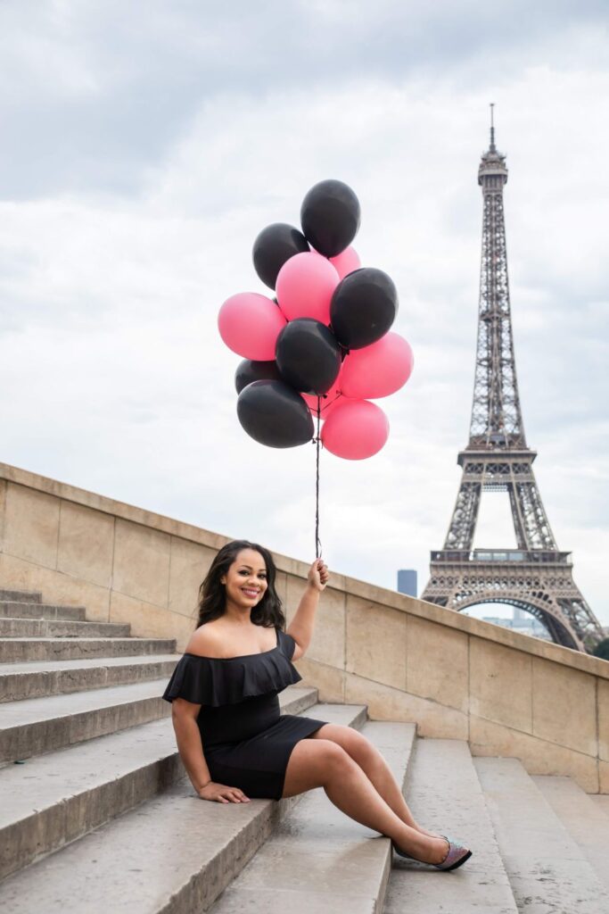 A joyful woman in a black off-the-shoulder dress sits on the steps at Trocadéro with a bouquet of pink and black balloons, the Eiffel Tower in the backdrop, capturing the essence of Parisian life at a beloved Paris instagram spots.