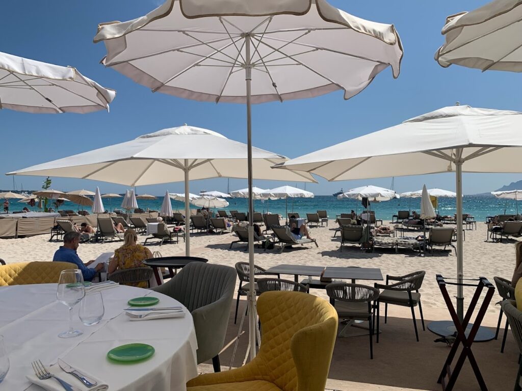 Diners relax under white parasols at Vegaluna Cannes Beach Clubs, with a view of the sandy beach and azure sea dotted with yachts.