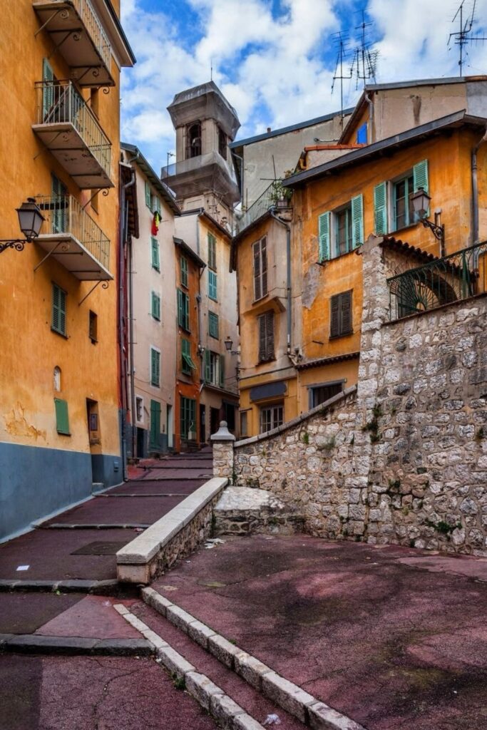 Narrow, sloping streets in the Vieille Ville (Old Town) of Nice, with a blend of colorful facades, green shutters, and historical charm under a soft blue sky. A perfect visual for a Nice travel guide, capturing the essence of the city's rich history and picturesque urban scenery.