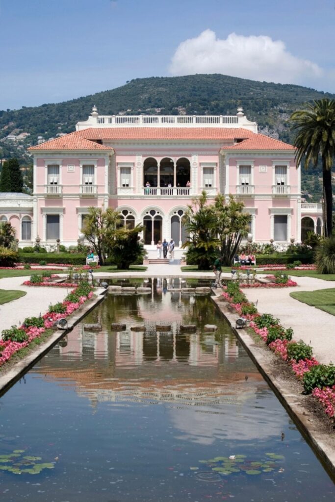 Symmetrical view of the grand Villa Ephrussi de Rothschild in Villefranche-sur-Mer, its pink walls and classical architecture reflecting in the still water of the ornamental garden pond. Flanked by lush greenery and blooming flowers, the estate offers a luxurious visual experience, perfect for those compiling a list of elegant things to do in Villefranche-sur-Mer.