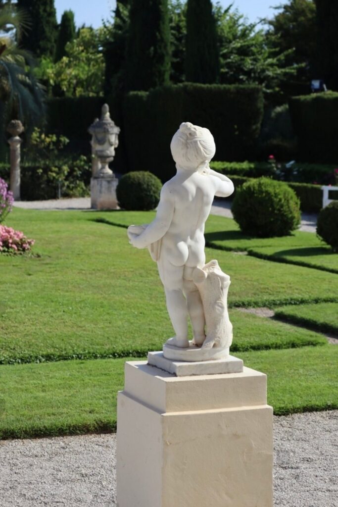 Elegant white marble statue of a child playing with a dog at the Villa Ephrussi de Rothschild in Villefranche-sur-Mer, set within a meticulously landscaped garden with geometric hedges and floral accents. This classical piece offers an enchanting detail to visitors exploring the art and gardens, a delightful activity among things to do in Villefranche-sur-Mer.