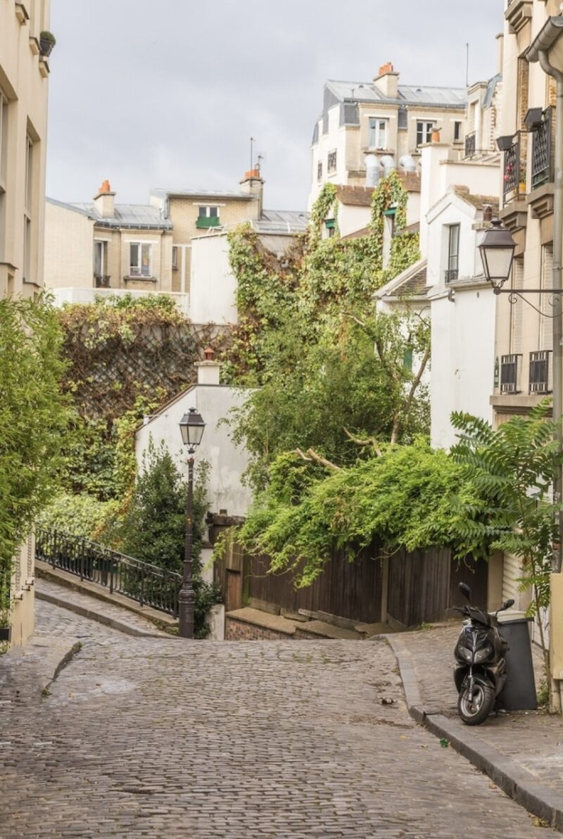 Cobblestone street of Villa Léandre in Paris with charming old buildings and lush greenery, with a solitary scooter parked beside a vintage lamppost.