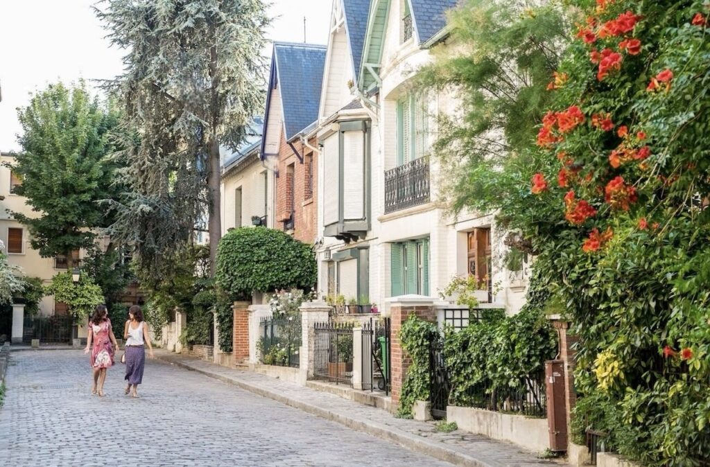 Two women walking down the picturesque Villa Léandre in Paris, with cobblestone streets and rows of elegant homes adorned with green shutters and vibrant flowers.