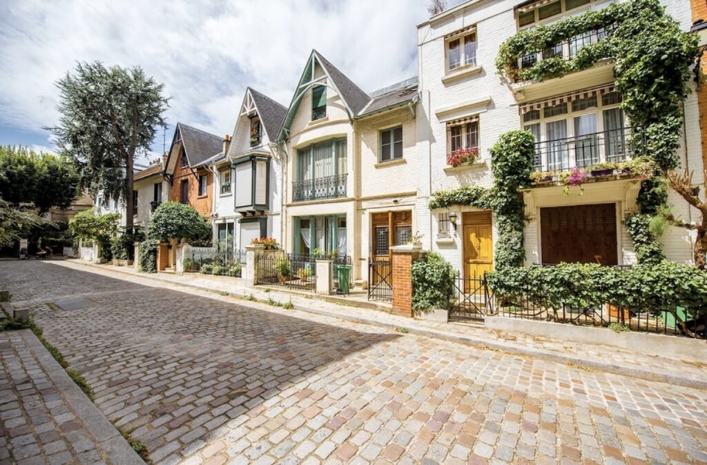Pristine cobblestone road leading through the tranquil Villa Léandre in Paris, flanked by elegant houses with colorful doors, ivy-draped walls, and blooming flower boxes.