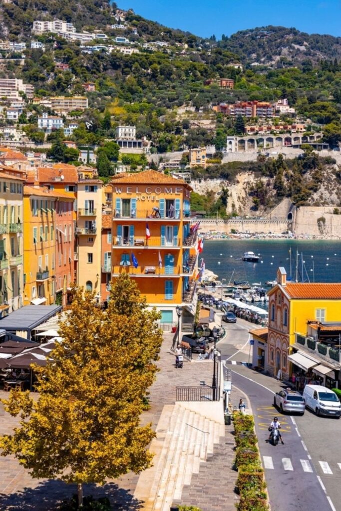 Overlooking the bustling quayside, the iconic 'Welcome Hotel' stands out with its terracotta façade among the vibrant buildings of Villefranche-sur-Mer. With the harbor, boats, and the hillside in view, this locale is a prime spot for visitors seeking the coastal charm and activities that are central to things to do in Villefranche sur Mer.
