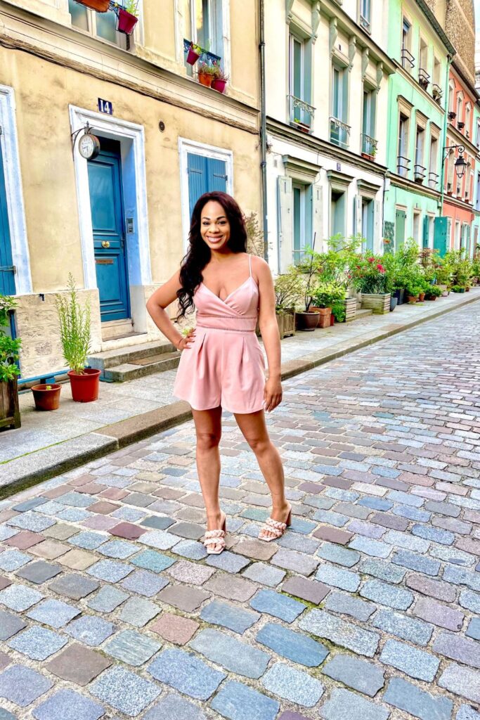 A woman smiles radiantly in a soft pink dress on Rue Crémieux, known for its colorful houses and cobblestone street, a picturesque and popular Paris instagram spots