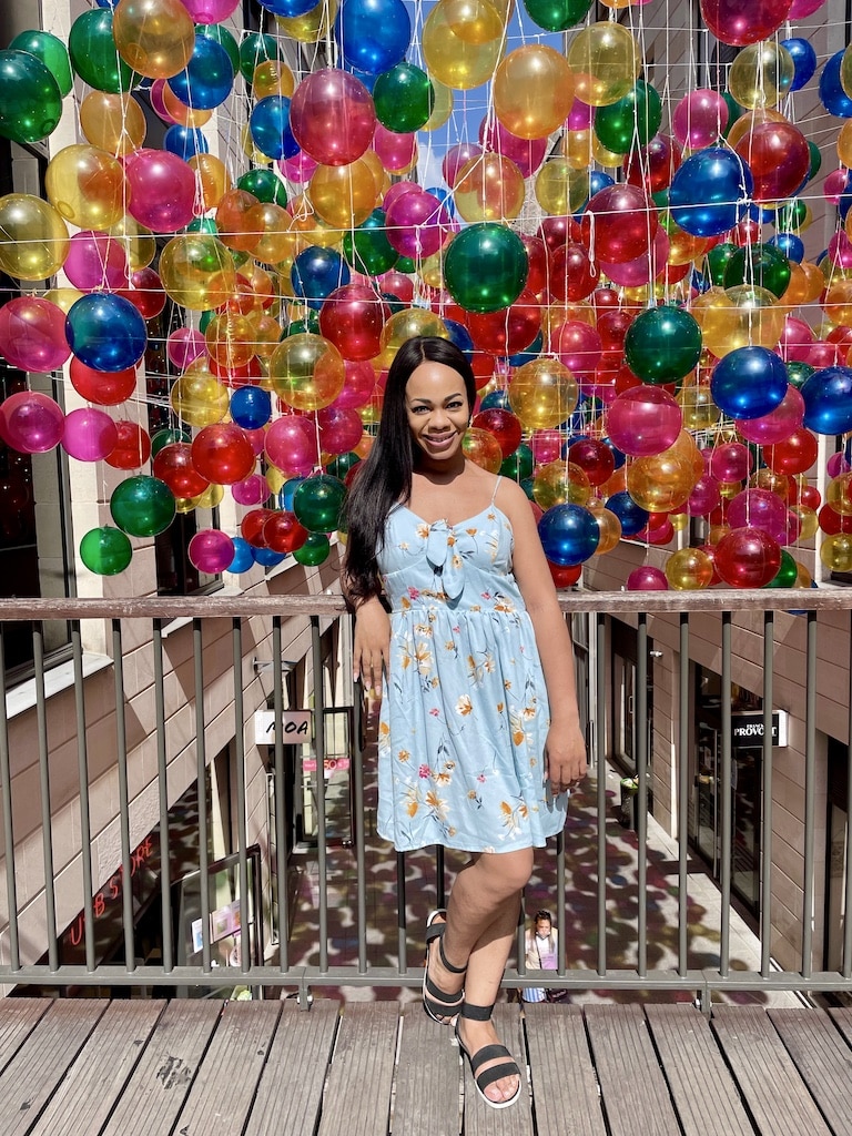 A woman smiles radiantly in a light blue summer dress adorned with a floral pattern, standing before a festive backdrop of multicolored balloons suspended in the air, capturing a moment of joy and celebration.
