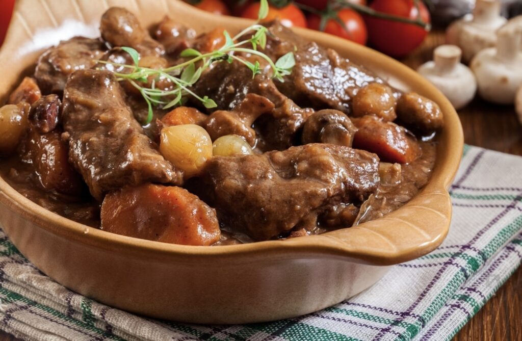 A hearty serving of Beef Bourguignon, a quintessential dish in the array of famous French foods, featuring tender beef chunks and vegetables in a rich gravy, garnished with fresh herbs, and set on a rustic tablecloth, ready to enjoy.