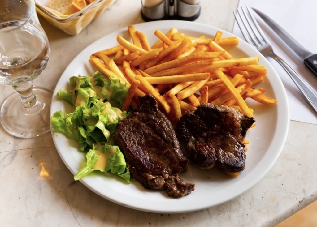 A plate of famous French foods featuring succulent beef steaks, crispy golden French fries, and a fresh green salad drizzled with dressing, accompanied by a glass of water on a marble table.