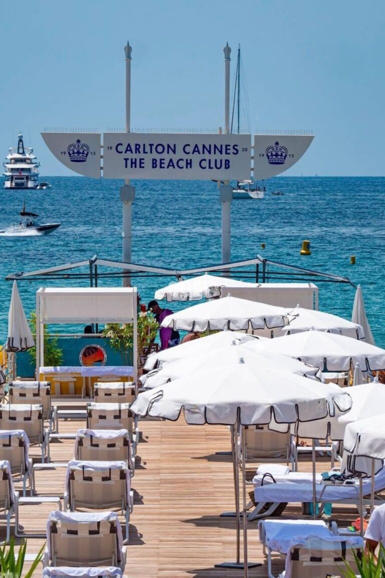 The Carlton Cannes Beach Club entrance, with a welcoming sign perched above a wooden walkway lined with white loungers and parasols, leading to the azure sea with luxury yachts and boats in the background. The scene captures the essence of exclusive Cannes beach clubs.