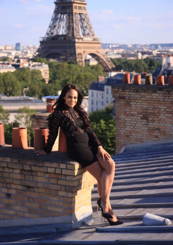 A woman in a chic black lace outfit sits on a rooftop ledge, smiling with the iconic Eiffel Tower in the background, capturing one of the best Paris Instagram spots.