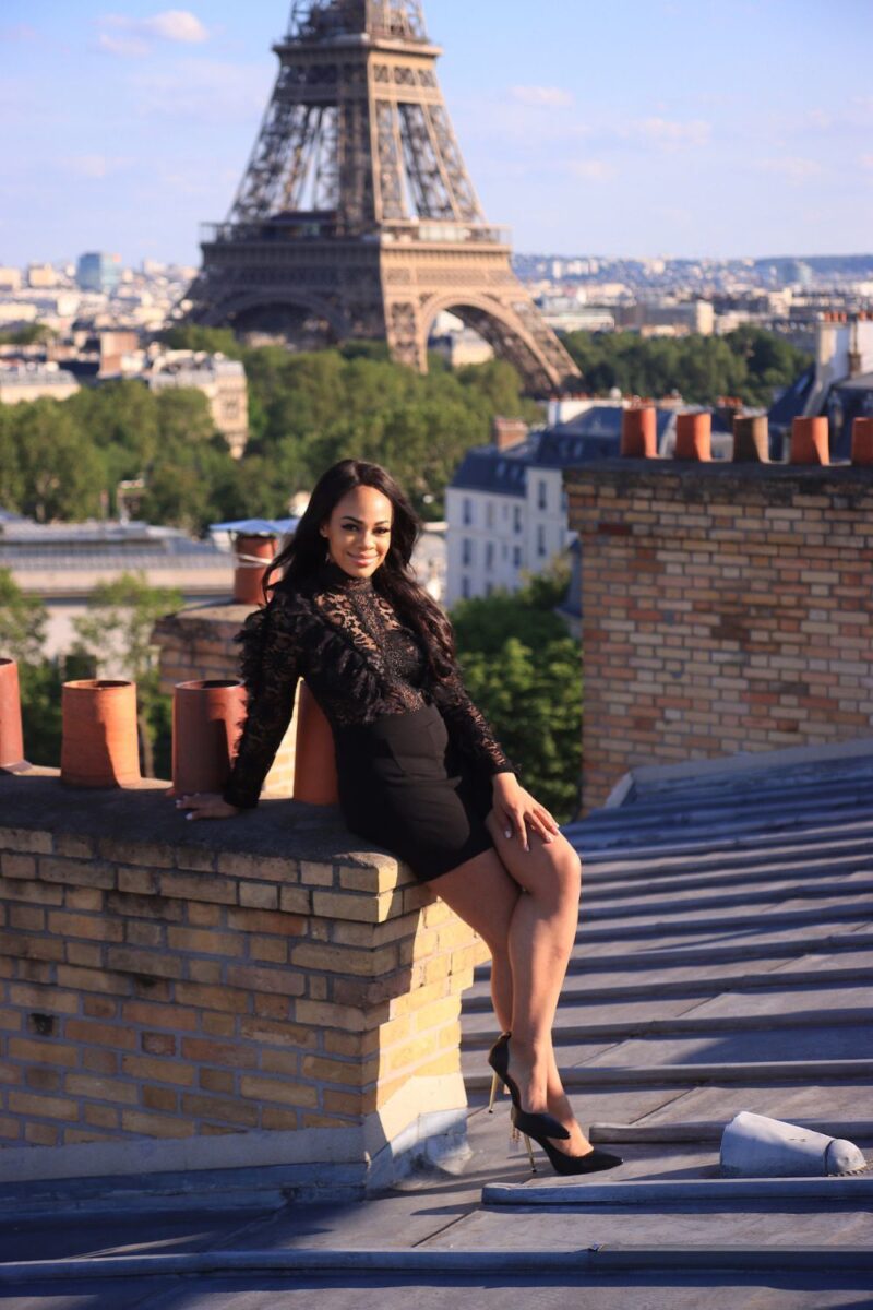 A woman in a chic black lace outfit sits on a rooftop ledge, smiling with the iconic Eiffel Tower in the background, capturing one of the best Paris Instagram spots.