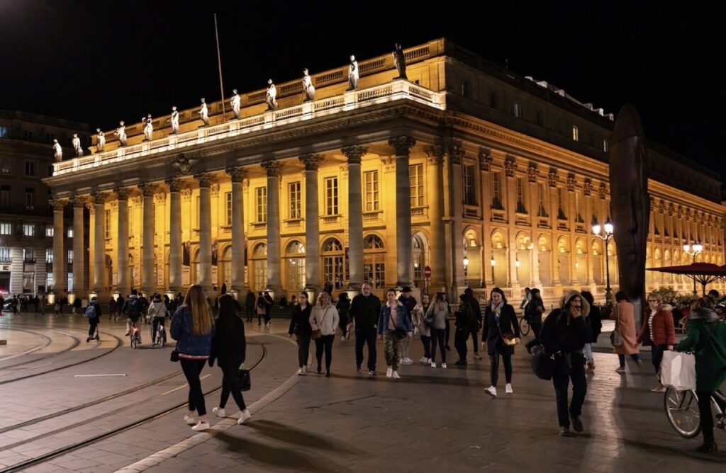 Night falls on Bordeaux as visitors and locals stroll in front of the illuminated Grand Théâtre, a premier cultural venue and architectural masterpiece, its facade warmly lit and statues standing guard above, a lively and elegant sight amongst the things to do in Bordeaux at night.