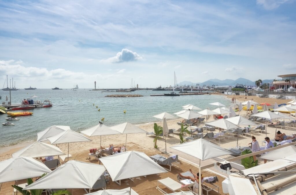 A lively day at a Cannes beach club captured from the terrace, with visitors relaxing under white parasols on sandy shores, pedal boats and yachts floating on the blue waters, and the scenic backdrop of Cannes' coastline and hills framing the tranquil Mediterranean setting.