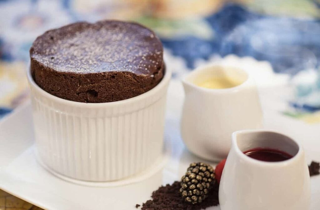 A perfectly risen chocolate soufflé, a gem among famous French foods, dusted with powdered sugar, accompanied by a smooth vanilla crème and a rich berry coulis, presented elegantly on a plate with a fresh blackberry garnish.