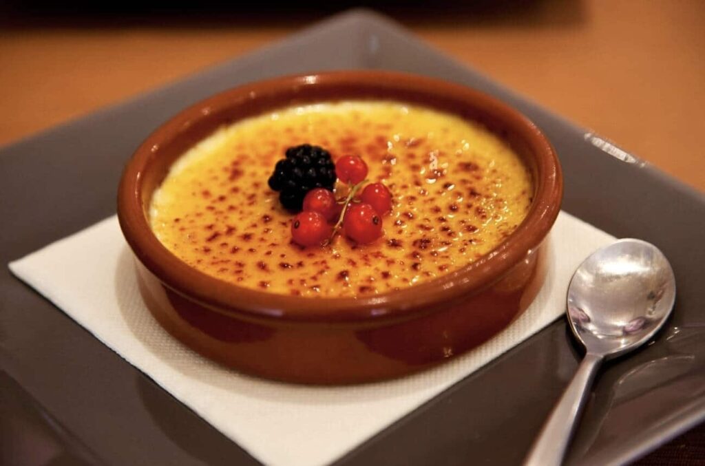 Crème brûlée, a decadent dessert known as one of the famous French foods, with a caramelized sugar crust and garnished with fresh blackberries and red currants, served in a classic ceramic dish alongside a small dessert spoon.