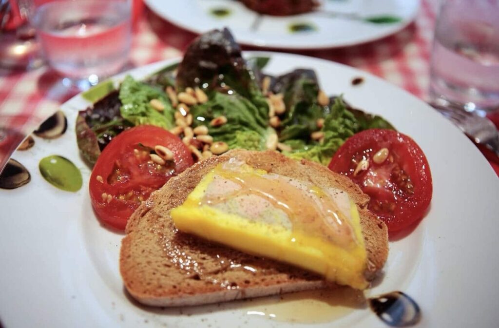 A traditional French dish featuring foie gras atop a toasted slice of bread, drizzled with a golden sauce, accompanied by a fresh salad with pine nuts and ripe tomato slices, capturing the essence of famous French foods.