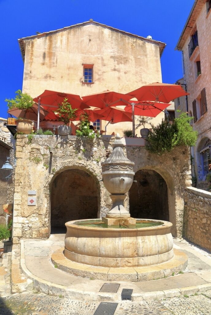An ancient stone fountain basks in the sunlight in Saint-Paul-de-Vence, a historic and must-visit Riviera cities. Overlooking the fountain are quaint buildings with red awnings, adding vibrant color to the rustic charm of this medieval town.