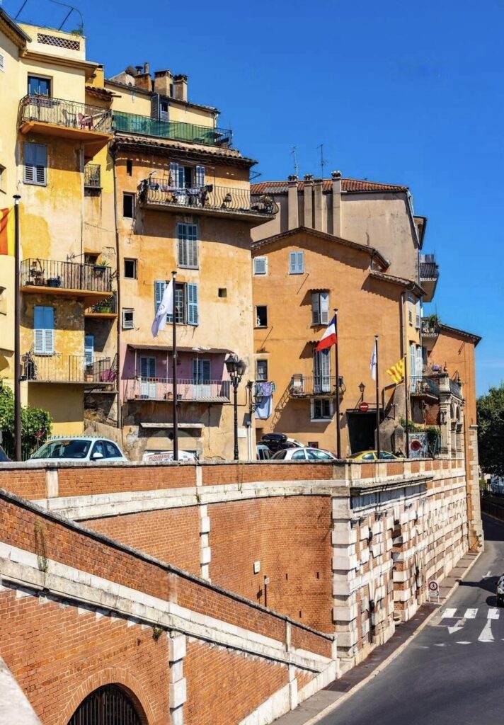 Warm-hued buildings with balconies adorned with flags line a sunlit street in Grasse, showcasing the city's vibrant charm. The historical architecture and clear blue skies make Grasse a picturesque, must-visit French Riviera cities.