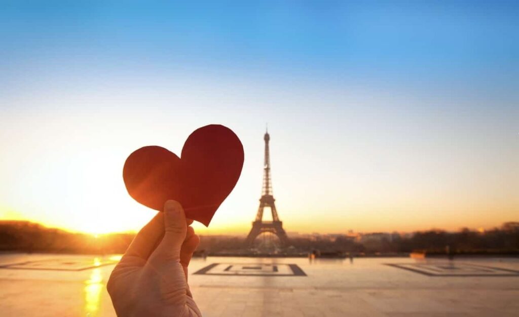 A hand holding a red heart-shaped cutout with the Eiffel Tower in the background during sunrise, capturing the romantic essence of Valentine's Day in Paris.