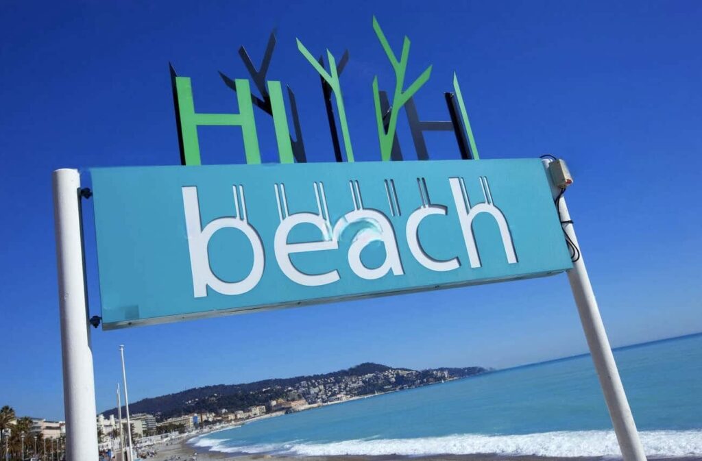 A vibrant sign with the text 'Hi beach' against a bright blue sky, overlooking the scenic coastline and sandy shores of Nice, ideal for promoting the best beach clubs in Nice.