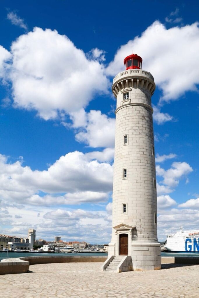The iconic lighthouse of Sète, standing tall against a backdrop of fluffy clouds and blue skies, overlooks the harbor, a notable landmark to see on a day trips from Montpellier.