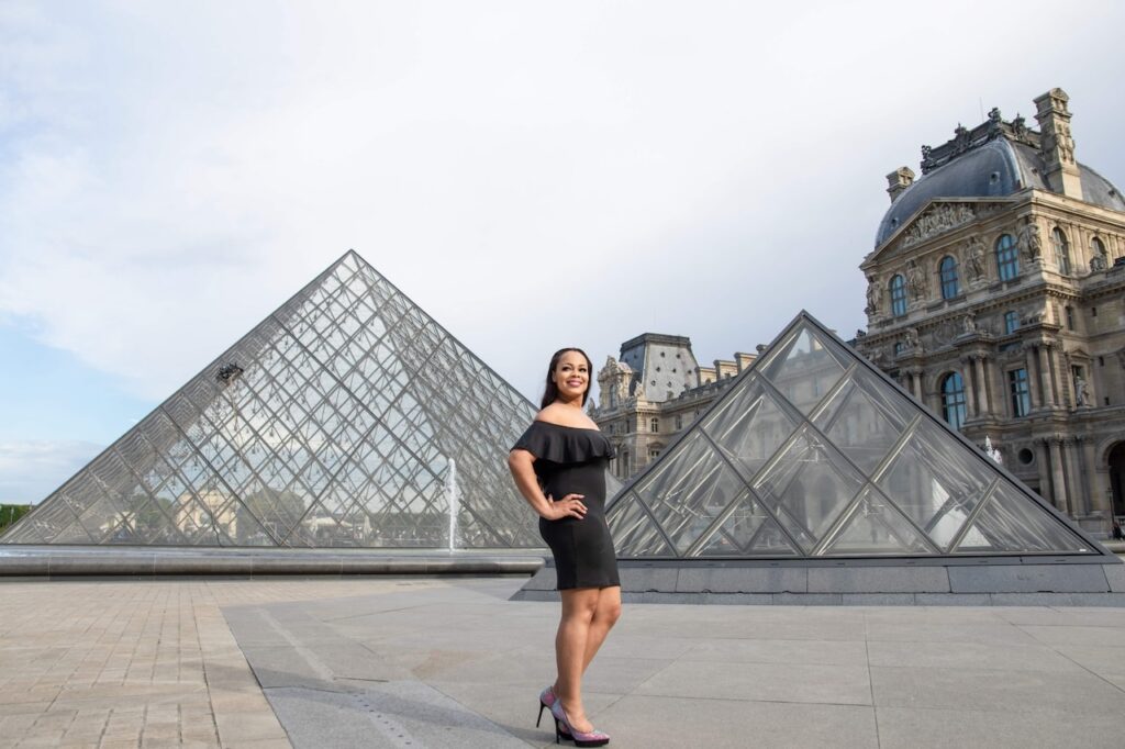 Confidently posing in a chic black dress, a woman stands in front of the Louvre Pyramid, an architectural marvel and a top Paris instagram spots, blending history with modern elegance.