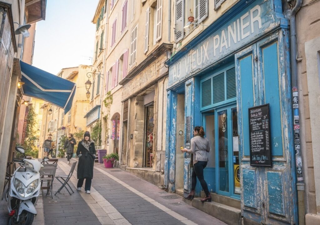 A quaint street scene in Le Panier, Marseille's oldest quarter, with people walking past the worn blue façade of "Au Vieux Panier." The rustic charm of the historic district is captured during One Day in Marseille.