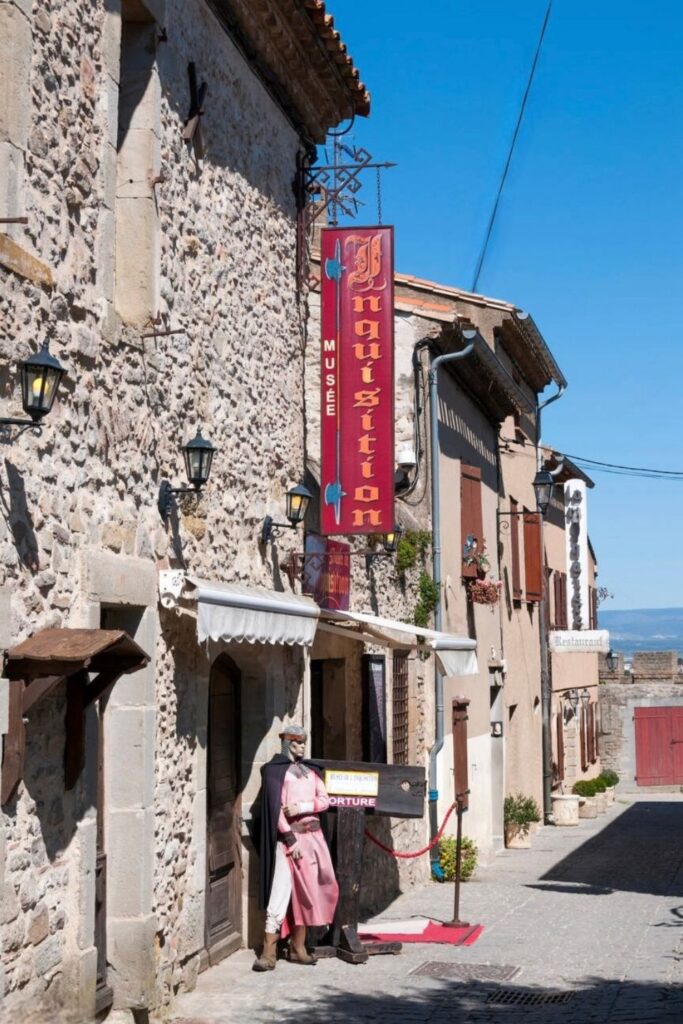 A quaint street in Carcassonne, France, featuring the stone facade of a museum with a colorful banner and a life-size figure in historical costume at the entrance. This museum adds a touch of intrigue to a day trips from Montpellier