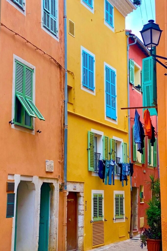 A stroll through the vibrant old streets of Villefranche-sur-Mer, where colorful laundry hangs above a quaint alleyway lined with traditional pastel-colored buildings and contrasting green shutters. This picturesque scene captures the charming daily life, an authentic slice of things to do in Villefranche-sur-Mer.