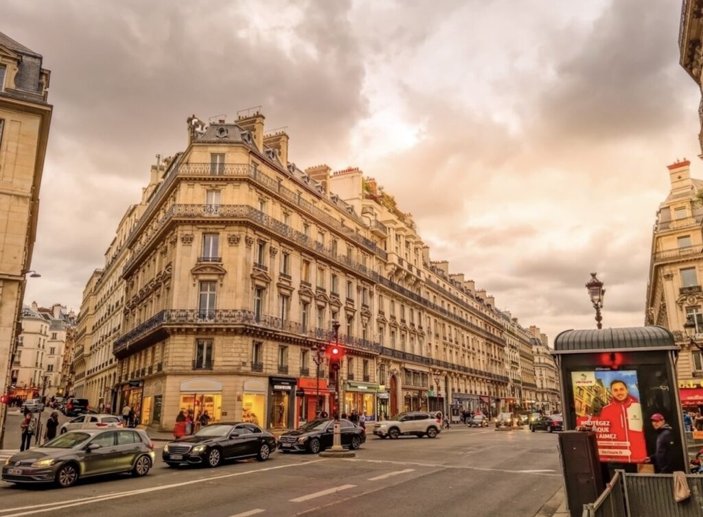 Twilight descends on a bustling corner of the best arrondissement to stay in Paris, showcasing elegant Haussmannian buildings and active city life with cars and pedestrians.