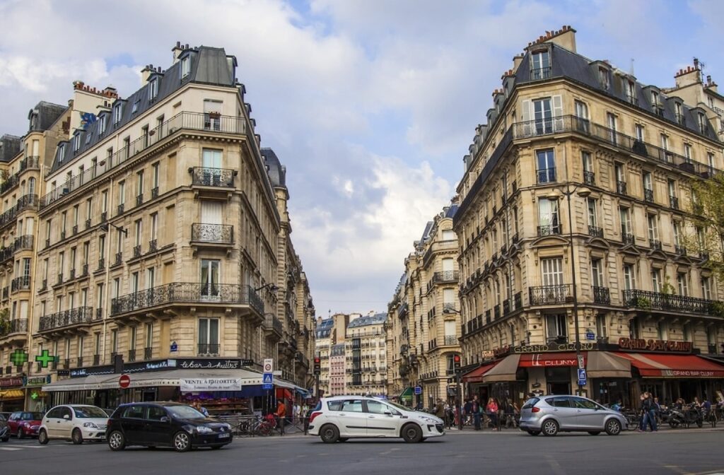 A lively intersection in the best arrondissement to stay in Paris, where traditional French architecture meets modern urban life with bustling cafés, busy traffic, and pedestrians mingling on the sidewalks.