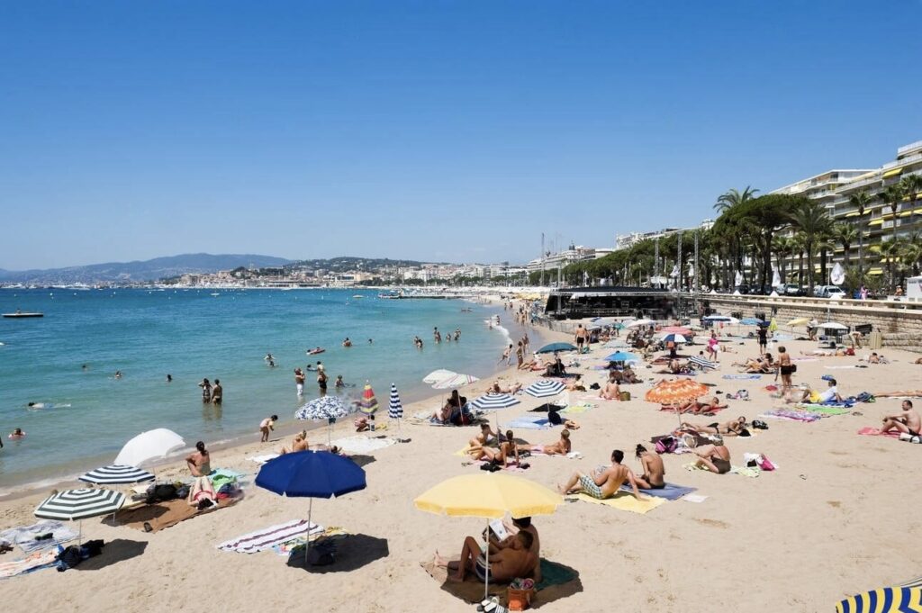 a bustling day at Plage de la Croisette in Cannes with sunbathers and swimmers enjoying the clear blue waters and sandy beach. Colorful umbrellas dot the shoreline against a backdrop of palm-lined promenades and the cityscape, capturing the vibrant atmosphere of Cannes beach clubs.