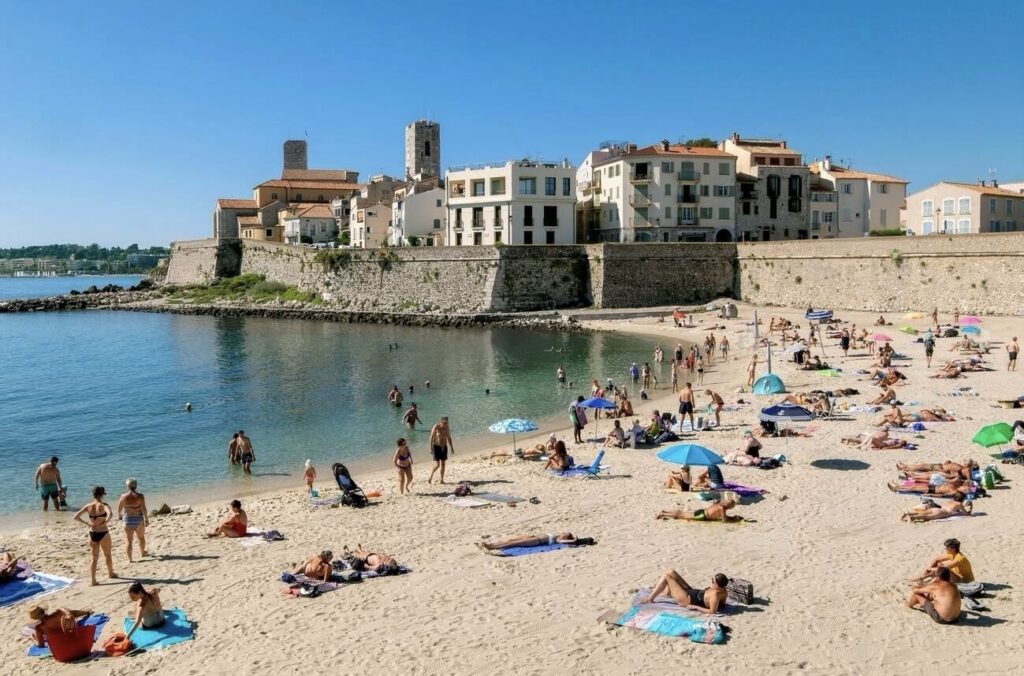Beachgoers enjoy the sun and sea at Plage de la Gravette in Antibes, a natural beach clubs in antibes setting, with people lounging on colorful towels and swimming in the calm bay, overlooked by the historic stone walls and architecture of Antibes.