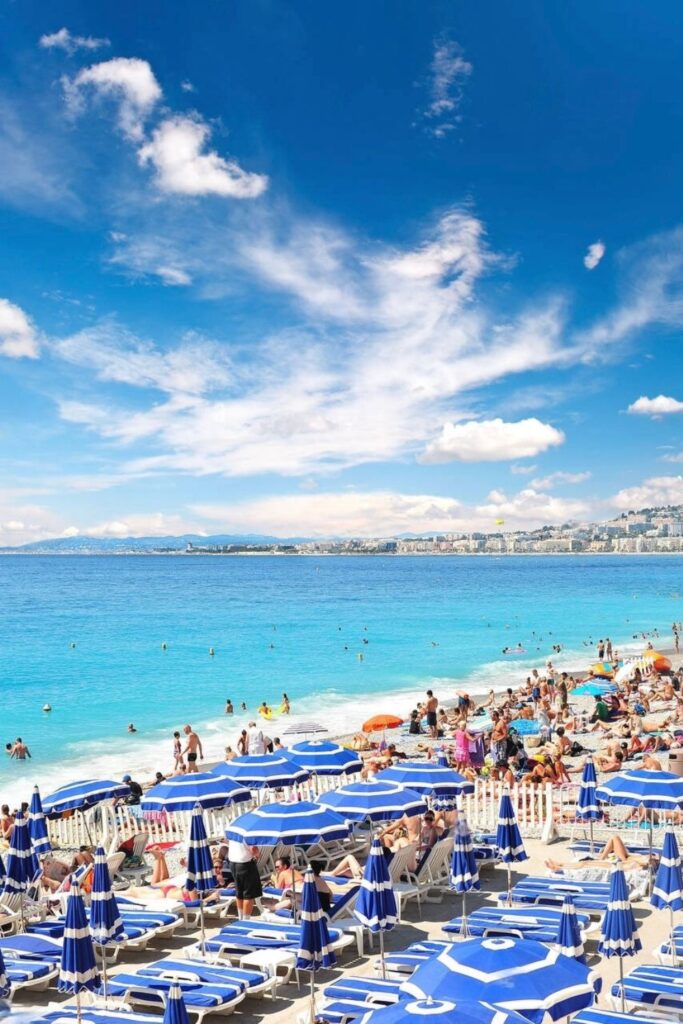 The bustling public beach of Nice, lined with blue and white striped umbrellas and sun loungers, as beachgoers enjoy the crystal clear waters of the Mediterranean Sea. A quintessential summer scene for a Nice travel guide, highlighting the city's lively beach culture against a backdrop of blue skies.