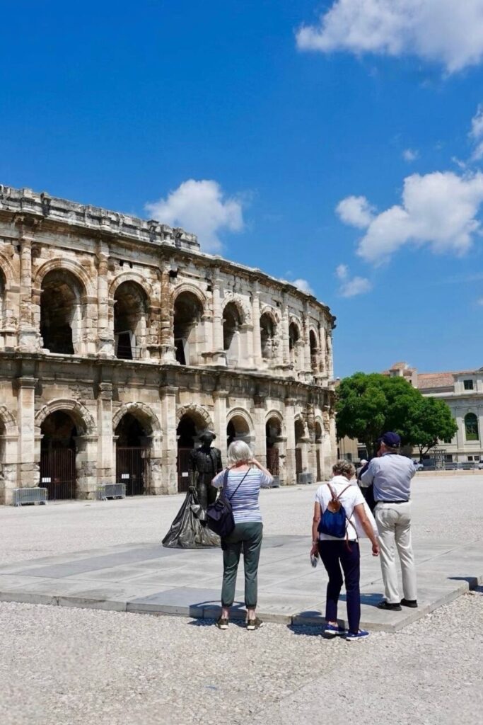 Tourists observe the ancient Roman amphitheater of Nîmes, France, under a bright blue sky dotted with clouds, capturing the grandeur of historical sites to be discovered on a day trips from Montpellier.