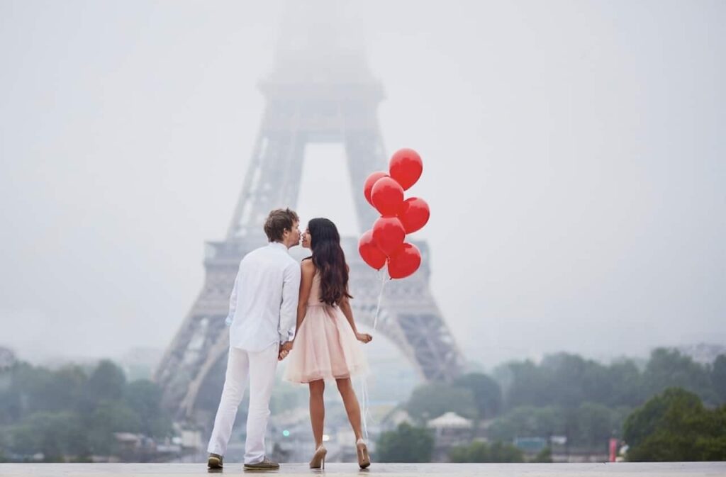 A romantic couple, dressed in white and pink, holds hands and shares a tender moment with a bouquet of red heart-shaped balloons, with the Eiffel Tower shrouded in mist in the background, evoking the spirit of Valentine's Day in Paris.