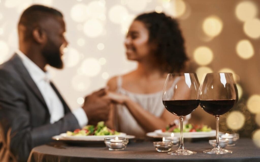 A couple enjoys a romantic dinner with wine glasses in the foreground and a backdrop of warm, glowing lights, symbolizing an intimate Valentine's Day in Paris.