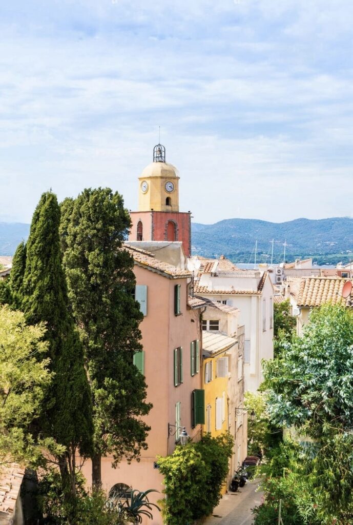 Alt text: "A view of Saint-Tropez's historic buildings with their pastel facades and green shutters, crowned by the iconic yellow bell tower. Tucked amidst lush trees, this scenic views captures the charming essence of one of the must-visit French Riviera cities.