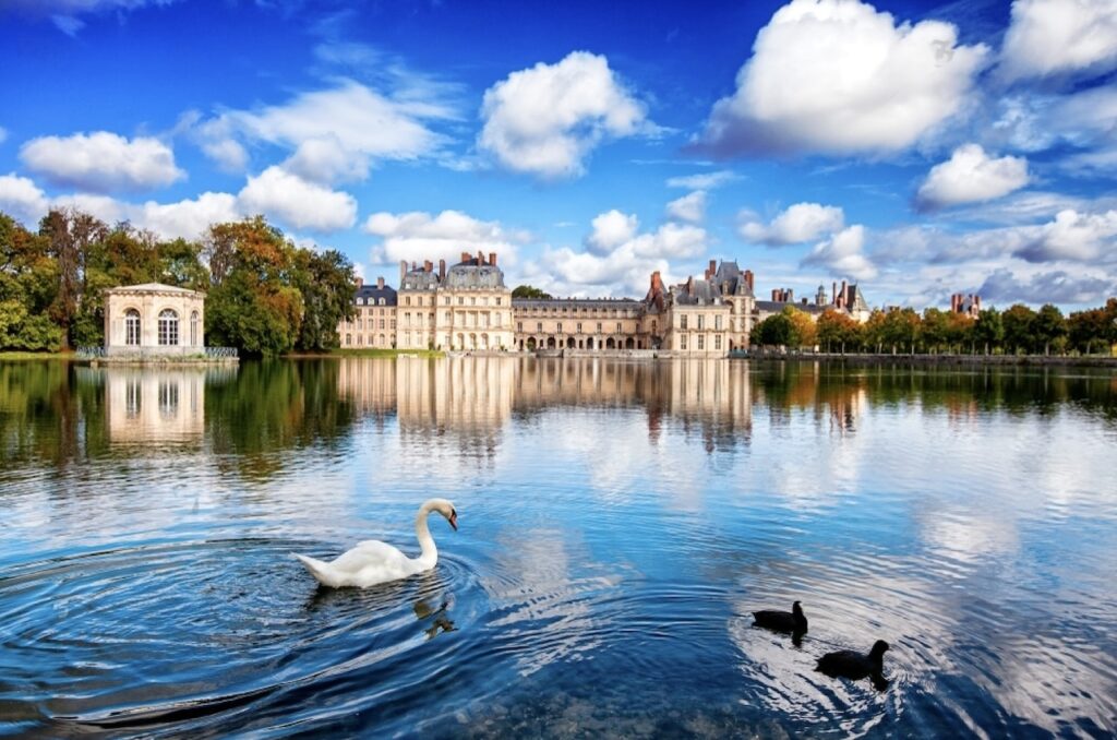 A swan and ducks glide peacefully on the water in front of the majestic Château de Fontainebleau under a vibrant blue sky, a serene destination for couples looking for things to do on Valentine's Day in Paris.