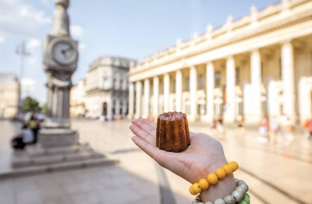 A hand presents a traditional Bordeaux canelé, a small French pastry with a caramelized crust and soft custard center, against the blurred backdrop of the Grand Théâtre in Bordeaux, evoking the city's culinary heritage as part of the things to do in Bordeaux.