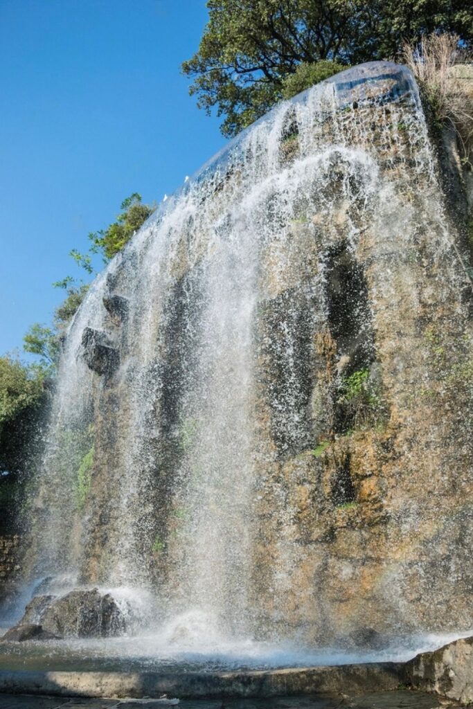 A majestic waterfall cascading down the rocky face of Colline du Château in Nice, surrounded by verdant foliage under a bright blue sky. A refreshing sight to feature in a Nice travel guide, highlighting the natural beauty within the urban landscape of the city.