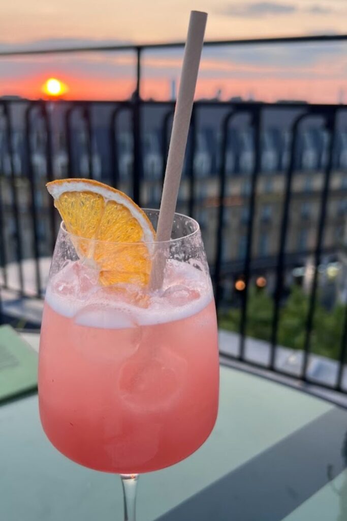 Alt text: A pink cocktail garnished with an orange slice and a paper straw sits on a table at a rooftop bar, with the sun setting over the Paris skyline in the background. Best rooftop bar in Paris.