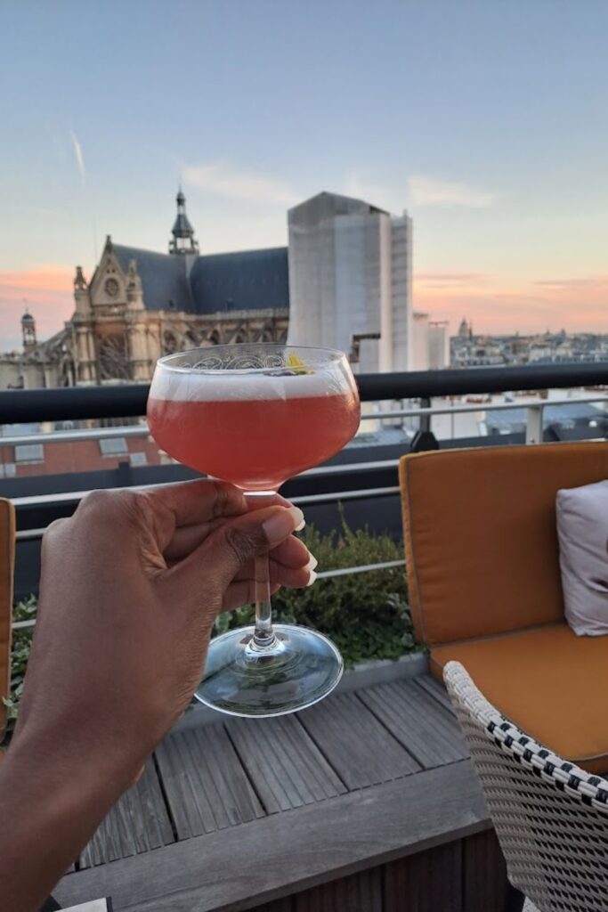 Alt text: A hand holding a pink cocktail in a coupe glass at a rooftop bar, with a view of a historic building and the Paris skyline during sunset. Best rooftop bar in Paris.