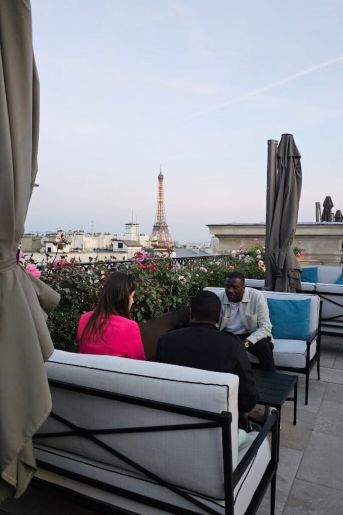 Alt text: A group of people sitting on cushioned benches at a rooftop bar, with the Eiffel Tower visible in the background against a clear evening sky. Best rooftop bar in Paris.