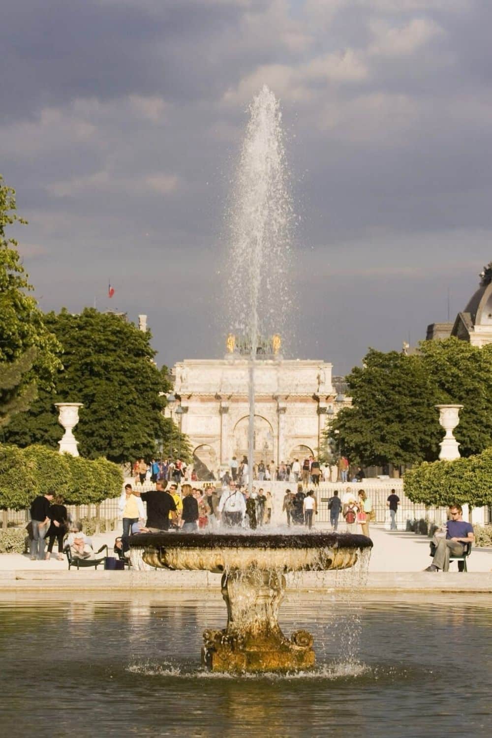 Fountain in the 1st arrondissement of Paris with water spraying into the air, surrounded by lush greenery and people walking and relaxing, with historic architecture in the background under a partly cloudy sky.