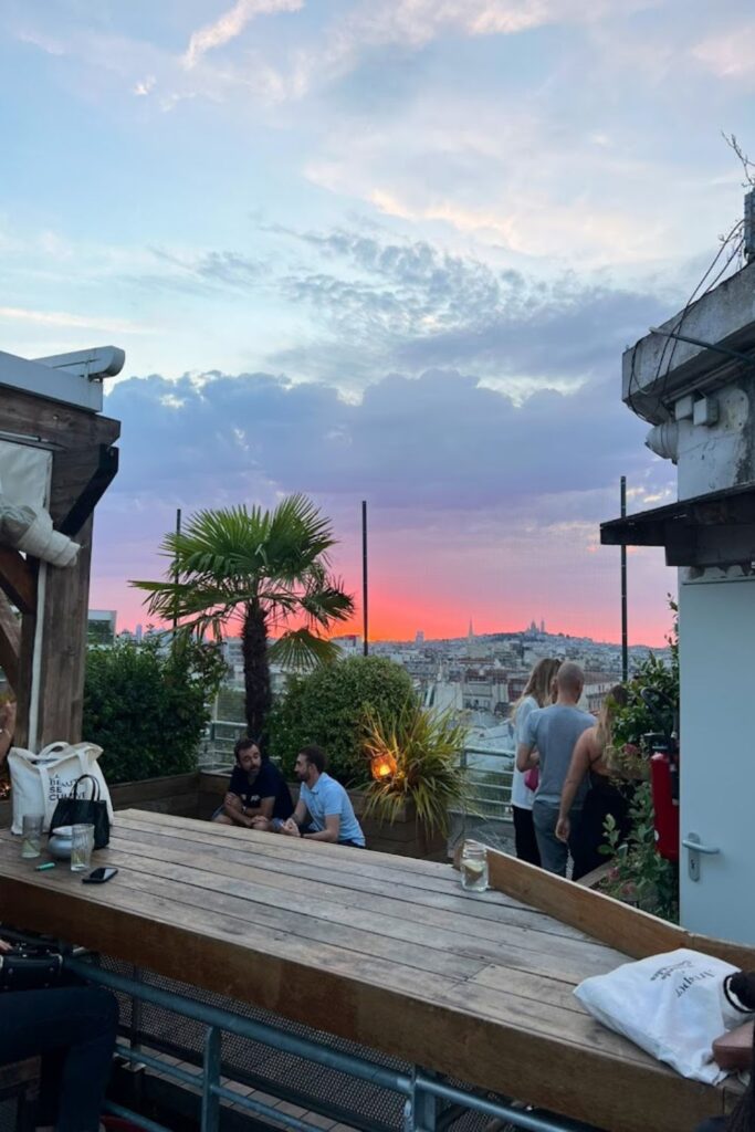 Alt text: People enjoying drinks and conversations at a rooftop bar during sunset, with vibrant sky colors and a view of Paris in the background. Best rooftop bar in Paris.